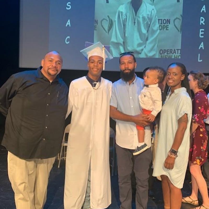 Class of 2019 graduate in white cap and gown with family in front of Operation New Hope backdrop