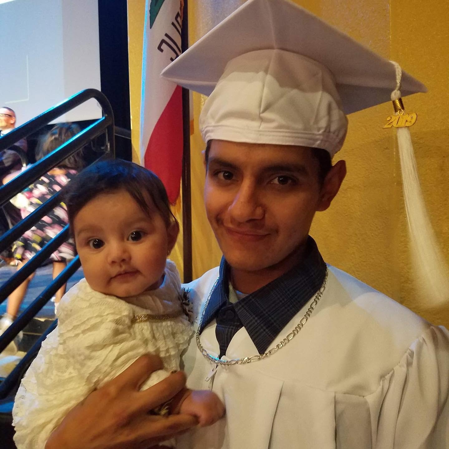 Class of 2019 graduate in white cap and gown holding a baby with California flag in background