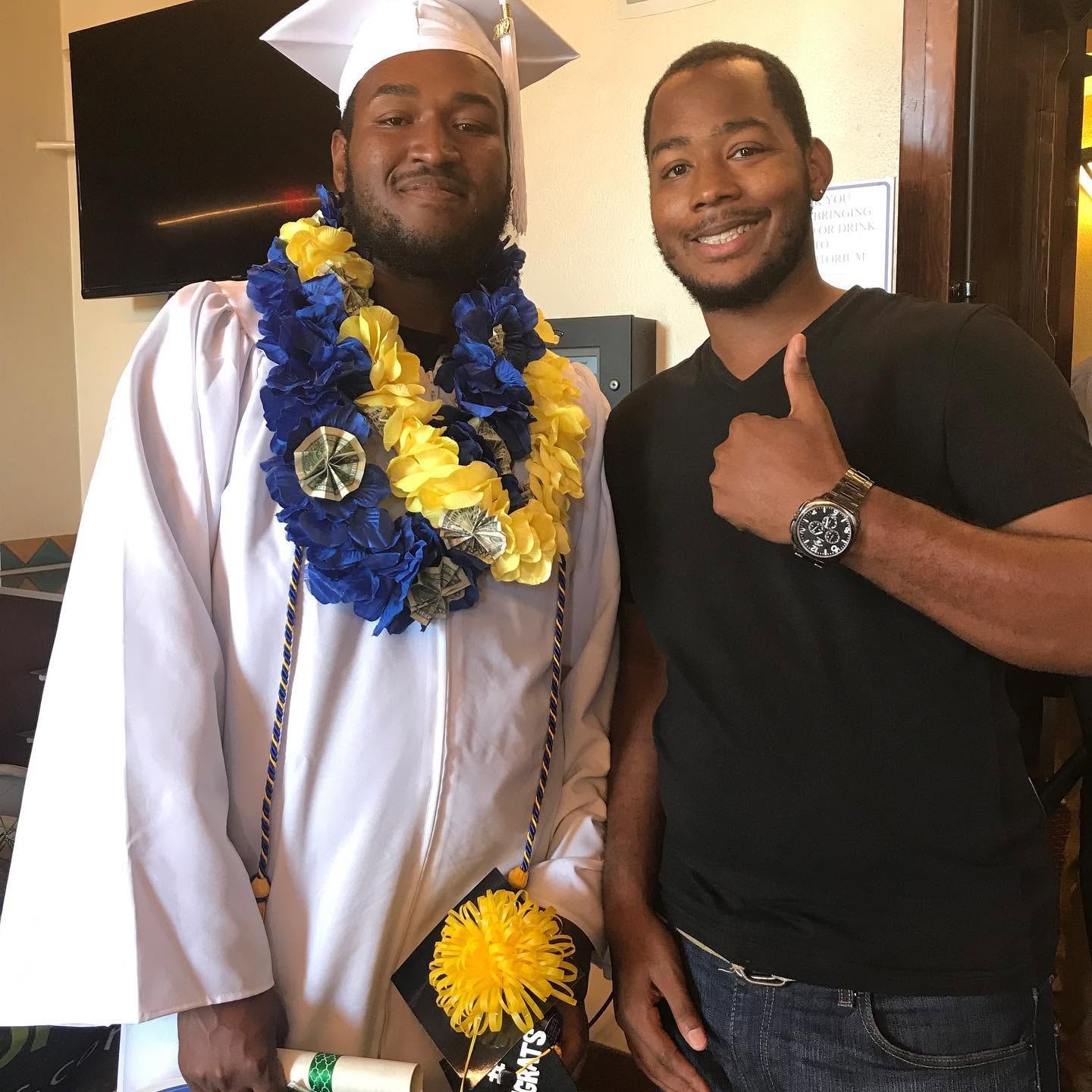 Class of 2019 graduate in white cap and gown wearing blue and yellow leis with supporter giving thumbs up