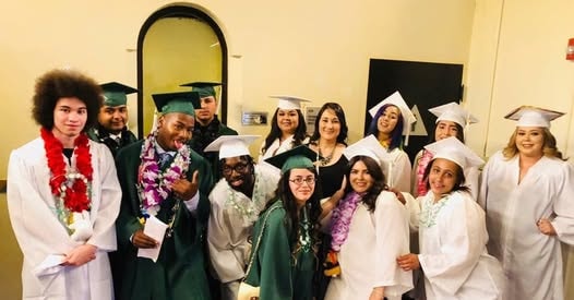 Large group of Class of 2019 graduates in green and white caps and gowns with leis celebrating indoors