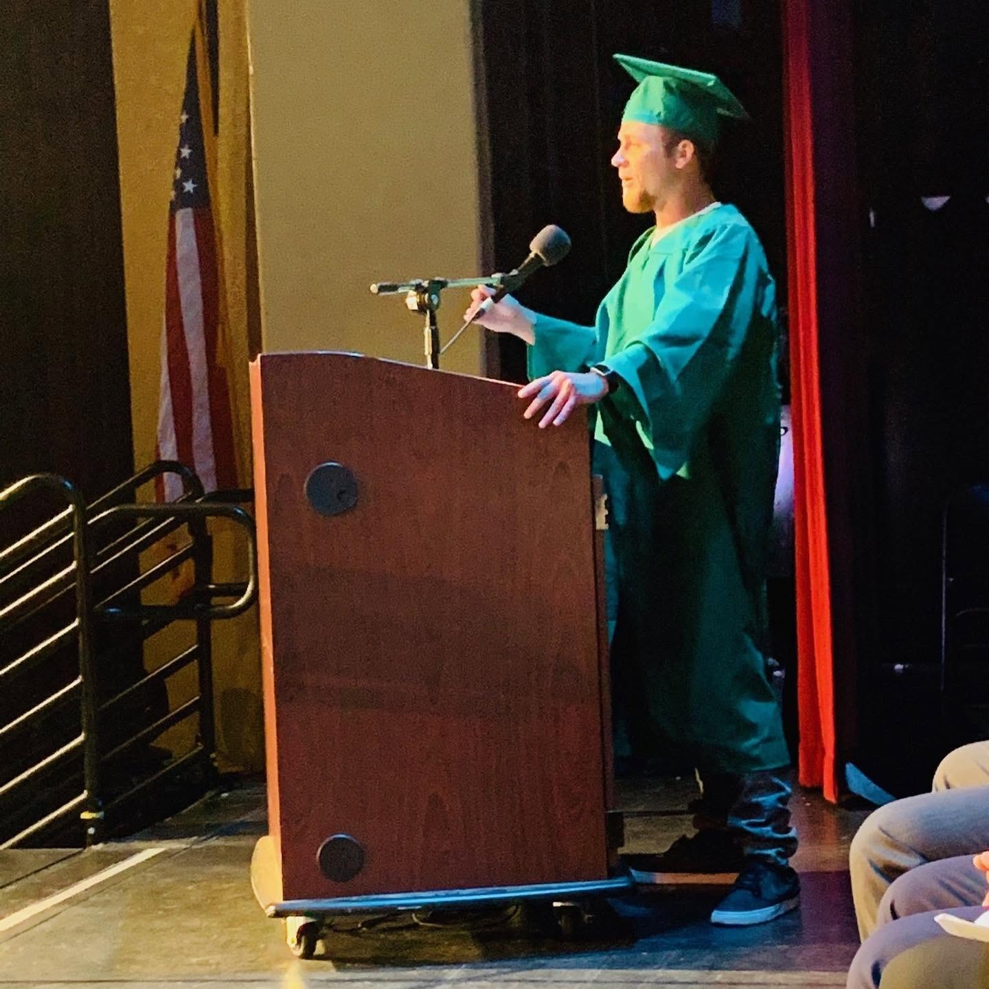 Zac, Class of 2019, delivering graduation speech at podium on stage in green cap and gown