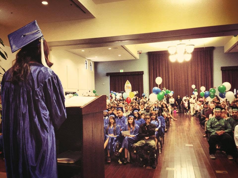 Graduate in blue cap and gown speaking at podium to packed graduation ceremony